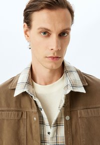 Brun manchesterjacka med två framfickor, lagd över en ljus rutad skjorta och en beige tröja med rund halsringning. Strukturerad och avslappnad.