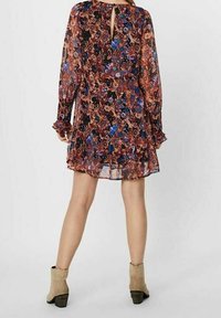 Šaty s květinovým vzorem, dlouhými průhlednými rukávy, výstřihem ve tvaru klíče na zádech a řasenými manžetami. Mají volný střih a barevný design na tmavém pozadí.