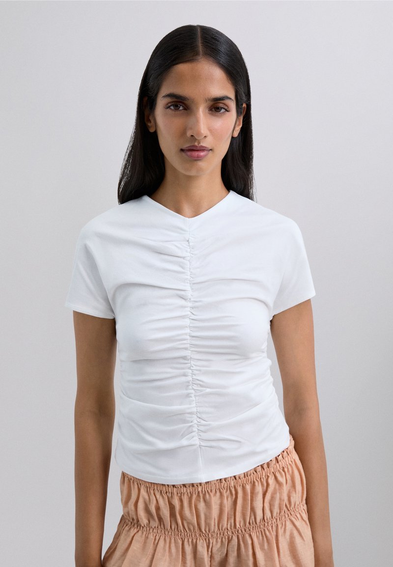 Mujer con pelo largo y oscuro, vistiendo una blusa blanca de mangas cortas con fruncidos y una falda corta de color melocotón, contra un fondo simple.