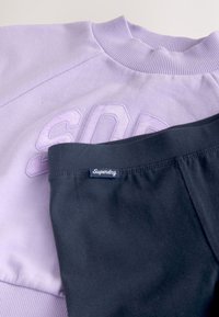 Lyse lilla sweatshirt med hævede bogstaver og sort Superdry undertøjs taljebånd placeret ovenpå.