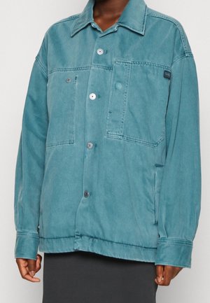 Personne portant une veste boutonnée surdimensionnée de couleur sarcelle avec de grandes poches frontales et des coutures visibles, associée à une jupe sombre.