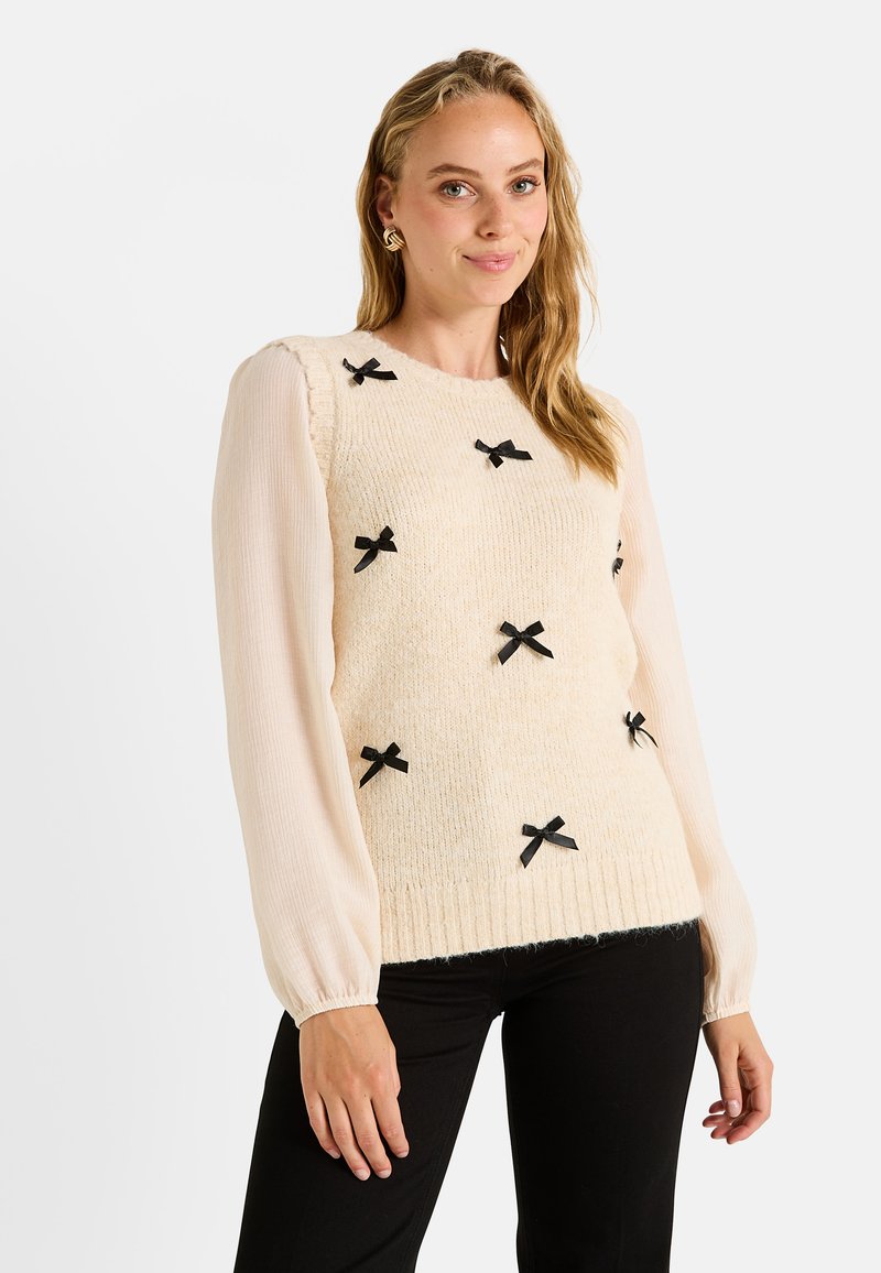 Pull en maille crème avec un devant texturé, des accents de nœud noir et des manches longues en voile. Ourlet côtelé, associé à un pantalon noir.