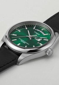 Zegarek Timex z zieloną tarczą, srebrnymi znacznikami godzin, wskazaniem daty na godz. 3 oraz czarnym skórzanym paskiem. Koperta ze stali nierdzewnej.