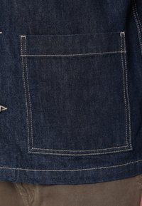 Chaqueta de mezclilla de color azul profundo con un bolsillo en el pecho bordeado con costuras en crema contrastantes. La tela tiene un acabado texturizado.