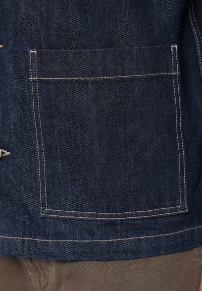 Veste en denim de couleur bleu profond, avec une poche poitrine soulignée de coutures crémeuses contrastées. Le tissu présente une finition texturée.