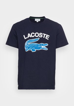 Marineblaues T-Shirt mit kurzen Ärmeln, weißem "LACOSTE"-Schriftzug und einem großen blauen Krokodil-Logo mittig auf der Vorderseite.