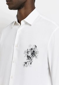 Camicia bianca in tessuto con una superficie liscia; presenta una stampa floreale nera e grigia nella zona sinistra del petto; design con bottoni.