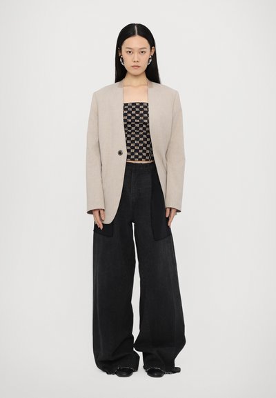 Beige skrædderjakke med en enkelt knap, lagt over en sort mønstret crop top, parret med brede sorte bukser og sorte sko.