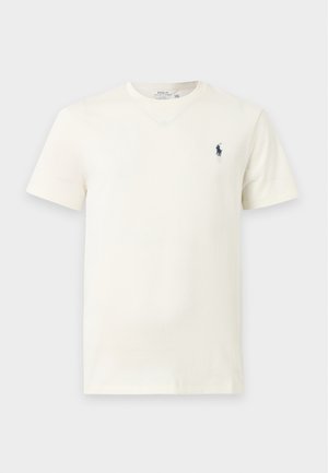 Νεαρός άνδρας όρθιος, φοράει κρεμ t-shirt, σκούρο παντελόνι, λευκά αθλητικά παπούτσια και καφέ καπέλο με μικρό λογότυπο τόσο στη μπλούζα όσο και στο καπέλο.