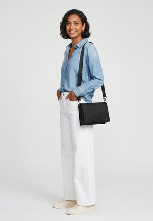 Kvinde i lyseblå denimskjorte, hvide bukser og beige sneakers står med hænderne i lommerne og bærer en sort crossbody taske.