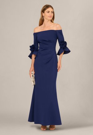 Kvinde iført en gulvlængde navy off-shoulder kjole med blomsterærmedetaljer, der holder en sølv clutch og står mod en ensfarvet baggrund.