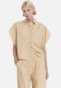Camisa de botones de manga corta beige con un bolsillo, que presenta mangas anchas y un corte holgado. El material parece suave y ligero.