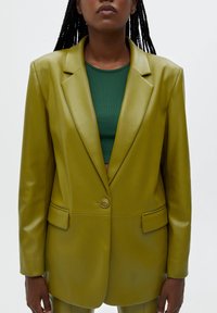 Olivgrön faux läder blazer med enstaka knappstängning, slagkrage och två framfickor med en slät yta.