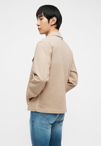 Νέος άνδρας με μαύρα μαλλιά, φοράει μπεζ μπουφάν και μπλε τζιν, στέκεται με τα χέρια στις τσέπες, στραμμένος πλαγίως, με φόντο απλό λευκό.