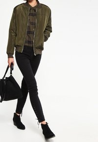 Olivgrüne Satinspringfieldjacke, karierter Hemd, schwarze Skinny-Jeans, schwarze Wildleder-Stiefeletten mit Schnürdetails und eine schwarze Tote-Bag.
