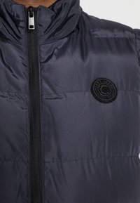 Marinblå vadderad väst med hög krage och frontdragkedja. Har en cirkulär svart logotyp på vänster bröst och en slät, blank yta.