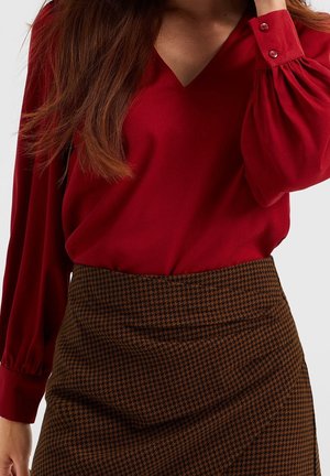 Vrouw draagt een rode V-hals blouse met lange, met knopen gesloten mouwen en een bruin-zwart pied-de-poule rokje, hand bij het haar.