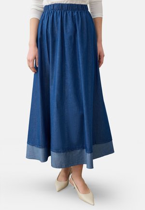 Kvinde iført et langt blåt denimskørt med en bred kant og elastisk talje, kombineret med cremefarvede slingback sko med spids snude.