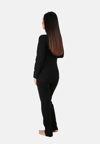 Top negro de manga larga y pantalones a juego, diseño ajustado, textura suave, usado por una persona de espaldas, descalzo, con el cabello largo visible.