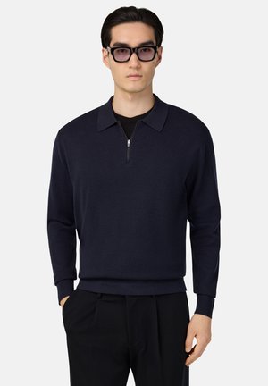 Dunkelblaues Strickpullover mit einem Reißverschluskragen, gerippten Bündchen und Saum. Der Stoff wirkt weich und strukturiert und wird mit schwarzen Hosen kombiniert.