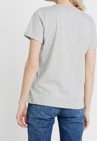 Šedé bavlněné tričko s krátkým rukávem, kulatým výstřihem a volným střihem, spárováno s modrými džínami. Zobrazeno ze zadní strany.