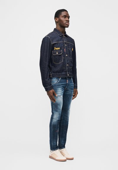 Hombre de pie vistiendo chaqueta de mezclilla oscura con bordado amarillo, vaqueros ajustados descoloridos con salpicaduras de pintura y zapatillas blancas sobre fondo liso.