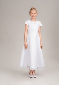 Weißes Kleid mit einem Spitzenoberteil und kurzen Ärmeln, fließendem Rock und transparentem Saum. Kombiniert mit silbernen Schuhen. Schlichtes und elegantes Design.