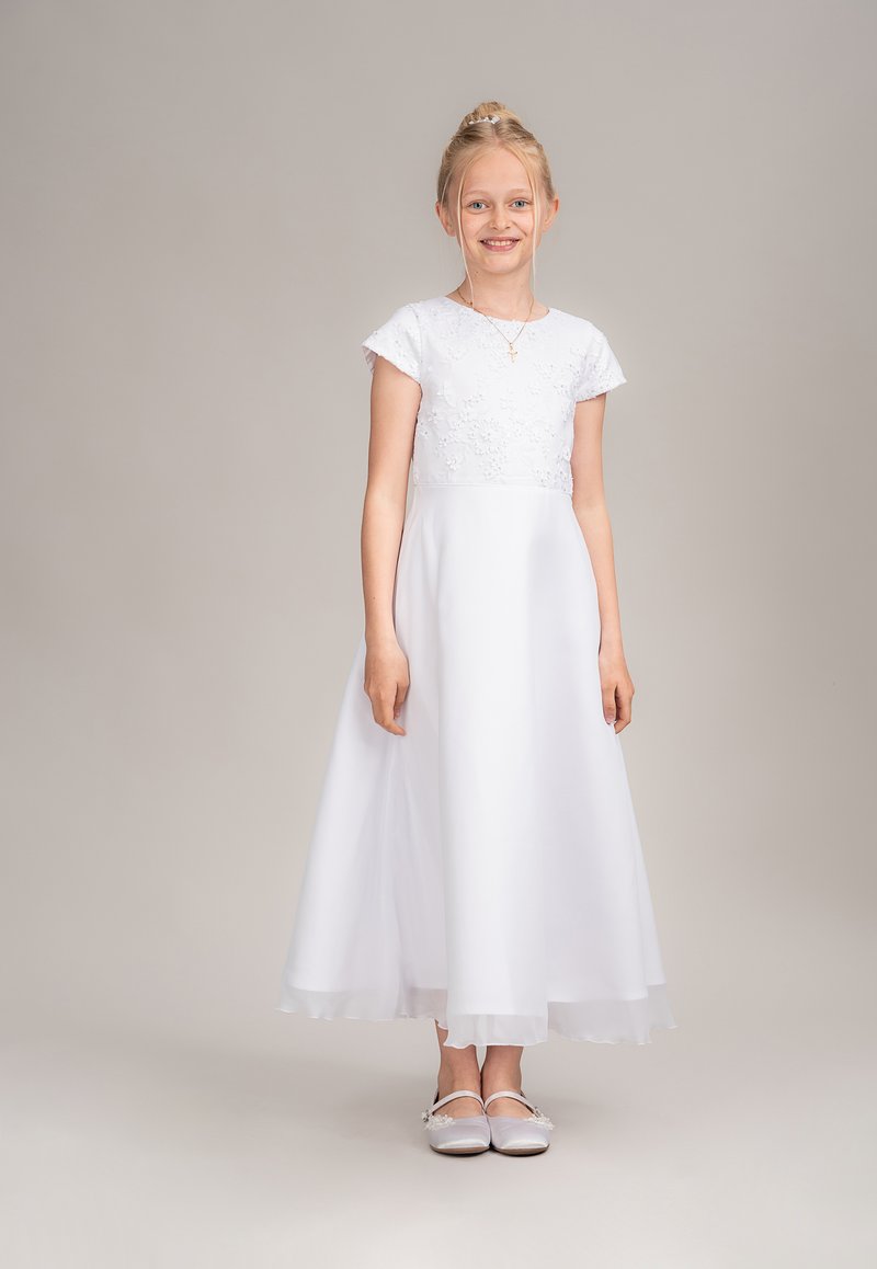 Weißes Kleid mit einem Spitzenoberteil und kurzen Ärmeln, fließendem Rock und transparentem Saum. Kombiniert mit silbernen Schuhen. Schlichtes und elegantes Design.