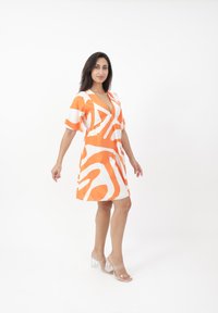 El vestido de patrón naranja y blanco presenta un escote en V profundo, mangas cortas y un ajuste relajado. Se lleva con sandalias claras de tacón bloque.