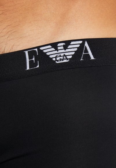 Chiloți negri dintr-un material neted și elastic. Prezintă o talie țesută cu un logo ce afișează un design cu o vultur și textul "E A."