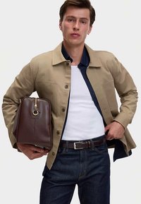 Beige katoenen jas met een kraag en knoopsluiting, gecombineerd met een wit overhemd. Donkerblauwe jeans en een bruine leren tas met gouden details.