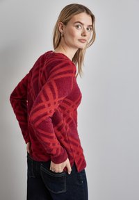 Rot gemusterter Pullover mit geometrischen Designs; aus strukturiertem Stoff gefertigt. Mit dunklen Jeans getragen, von der Seite gezeigt mit den Händen in den Taschen.