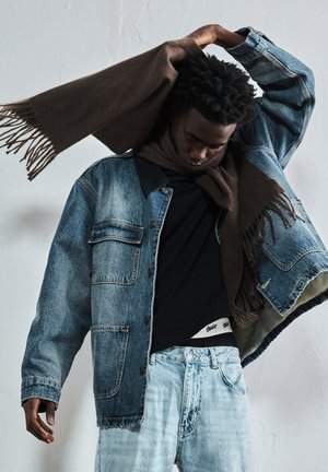 Man med ljusa denimjeans, en denimjacka, svart skjorta och en brun scarf runt halsen mot en enfärgad vägg.