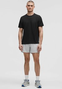 Schwarzes T-Shirt mit kurzen Ärmeln, kombiniert mit hellgrauen Shorts. Die Shorts haben ein strukturiertes Muster. Das Modell trägt graue Sneaker mit blauen Akzenten.