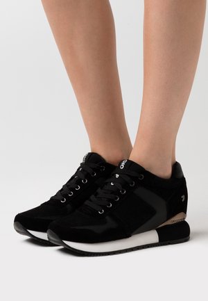 Pair of black lace-up sneakers worn on bare feet, featuring white soles and subtle metallic accents on the heel.