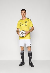 Yellow football jersey with black stripes, featuring a graphic design and logo. Paired with white shorts, black socks, and cleats, holding a soccer ball.