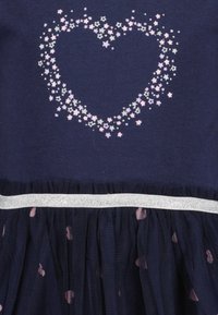 Marindress med en texturerad tyllkjol. Har ett hjärta gjort av pastellfärgade stjärnor på bodicen och ett silverband i midjan.