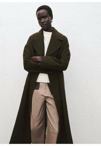 Manteau en laine vert olive avec de larges revers, superposé sur un pull blanc et un pantalon beige taille haute, présentant une texture lisse et une coupe ajustée.