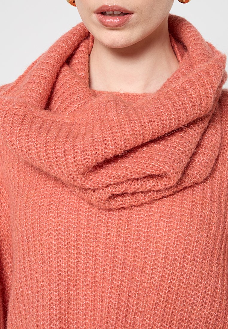 Maglione morbido in maglia di colore pesco con un ampio collo a scialle drappeggiato. La texture appare pelosa, con una trama allentata e un motivo a coste.