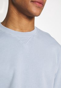 Světle modrá mikina s žebrovaným kulatým výstřihem, která má na výstřihu šikmé švy do tvaru V a jemnou, hladkou texturu.