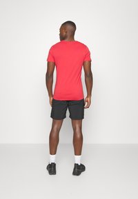 Red short-sleeve t-shirt, relaxed fit, paired with black shorts. Model wearing black sneakers and white socks, seen from the back.