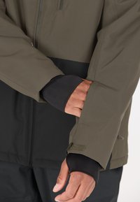 Wasserabweisende Jacke mit einem olivgrünen oberen Teil und einem schwarzen unteren Abschnitt. Verfügt über ein strukturiertes Gewebe, elastische Bündchen und Klettverschlussanpassungen.