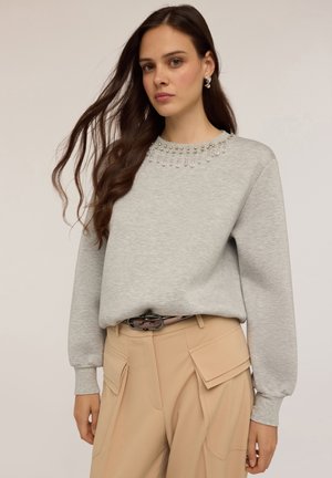 Felpa grigia con un colletto decorativo in perline. Maniche lunghe e vestibilità rilassata. Abbinata a pantaloni beige con molte tasche.