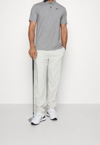Grau Polo-Shirt mit einem Kragen, weiße leichte Hose und weiße Sneakers mit schwarzen Akzenten. Ein Golfclub wird in der linken Hand gehalten.