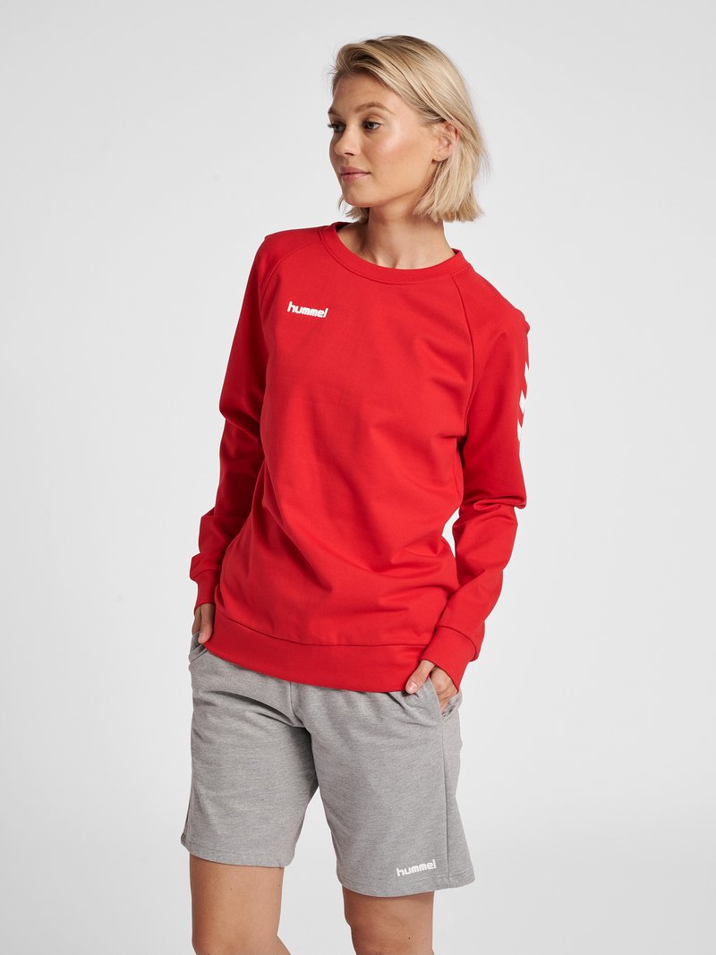 Femme aux cheveux blonds courts portant un sweat rouge hummel et un short gris hummel, mains dans les poches, regardant vers sa gauche.