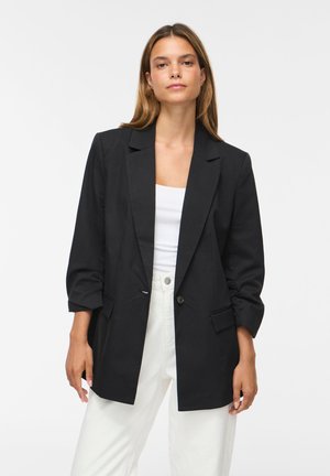 Ung kvinde med langt brunt hår iført en sort blazer over en hvid top og hvide bukser, stående mod en ensfarvet lys baggrund.
