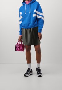 Felpa blu con strisce bianche, gonna in eco-pelle verde, borsa rosa e scarpe da ginnastica nere con calze bianche. Outfit casual e sportivo.