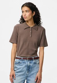 Brūns polo krekls, kas izgatavots no mīksta auduma, ar klasisku apkakli un trīs pogām; savienots ar gaiši ziliem džinsu biksēm un sapītu jostu.