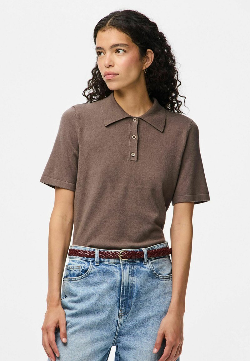 Brūns polo krekls, kas izgatavots no mīksta auduma, ar klasisku apkakli un trīs pogām; savienots ar gaiši ziliem džinsu biksēm un sapītu jostu.