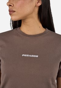 T-shirt corta marrone in cotone con colletto a girocollo, decorata con testo ricamato bianco "PEGADOR" sul petto. Texture liscia.